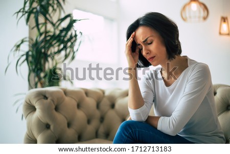 Headache. Close-up photo of a young woman, who is sitting on a sofa with her eyes closed, touching her head while suffering from a migraine.