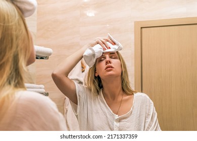 Headache, Migraine. Portrait Reflection In Mirror Of Young Girl Making Cold Compress With Towel On Her Head. Woman Standing In Front Of Mirror In Bathroom Throwing Her Head Back And Closing Eyes