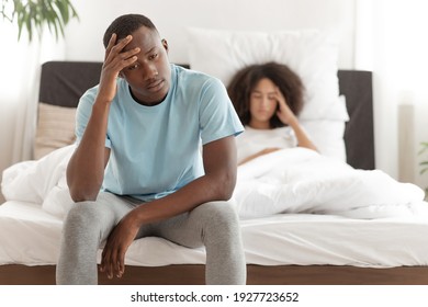 Headache, problems with potency and male power, quarrel and scandals. Upset millennial african american guy sits on bed and pressed his hand to forehead, upset wife in modern bedroom, copy space