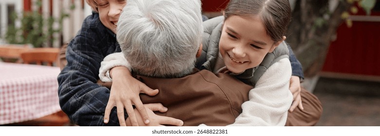 Header of elderly man embracing two smiling children outdoors in village setting, children hugging from both sides, faces joyful, intergenerational family bonding moment