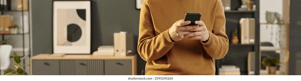 Header image of smiling young man using phone while standing in office and checking messages via internet, copy space