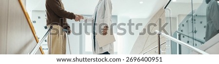 Header of middle aged man with disability using crutches shaking hands with young adult female doctor in white lab coat, standing in modern medical facility near staircase