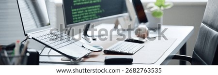 Header of modern office workspace featuring desktop computer with code on screen, wireless keyboard, mouse, laptop on stand, smartphone and accessories arranged on desk in bright environment