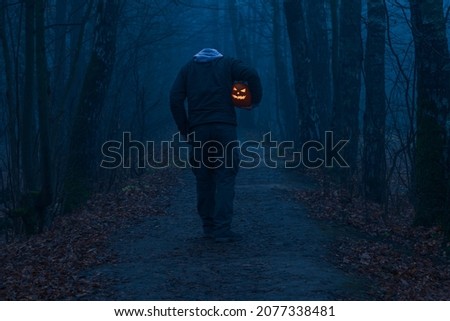 Headless man holds Jack O'Lantern Halloween pumpkin head under his arm and walks on countryside road in autumn foggy forest. Carved face on the pumpkin glows orange. Halloween costume theme.