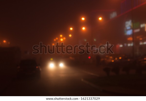 Headlights of car driving in fog at night in the\
city. lamp post lanterns, mist fog and street lights. Out of focus.\
Space for text.
