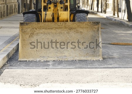 Head-on view of yellow wheel loader stationed on stripped asphalt corridor during municipal roadway intervention. Depicts mechanized contractor readiness, heavy equipment deployment, and active urban surface rehabilitation.