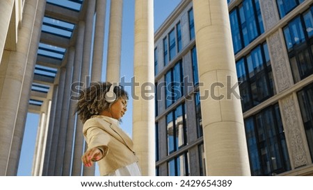 With headphones on and a radiant smile, a young professional woman dances freely in an urban space, her dynamic movement contrasting with the static lines of the surrounding architecture.