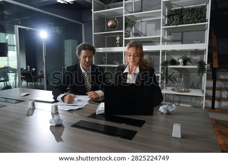 Heads angled toward the same document, the man and woman reconcile their notes at the night desk, teamwork closing out a demanding day in a bright modern coworking office.
