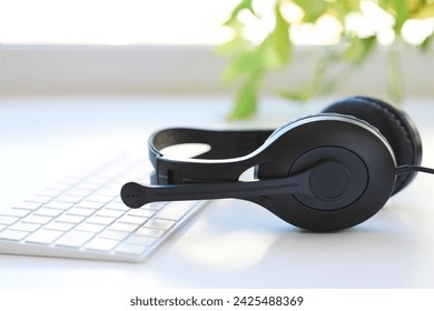 Headset with microphone and keyboard, placed on a white desk. Bright background light streaming in from the window. Close-up view. Remote work or learning concept.
