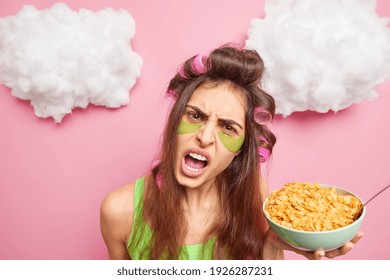 Headshot Of Angry Displeased Dark Haired Woman Exclaims Loudly Keeps Mouth Opened Reproaches You Because Of Sleep Interruption Has Breakfast Holds Bowl Of Cornflakes Makes Hairstyle With Curlers