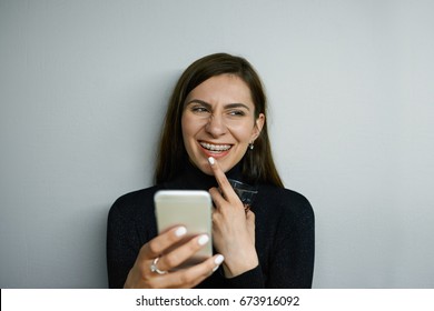 Headshot Of Attractive Cheerful Young Female With Long Dark Hair And Braces Texting Messages Using Online Messenger On Her Mobile Phone, Having Flirty Look, Touching Lips And Smiling Broadly