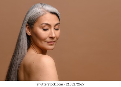 Headshot Of Attractive Middle-aged Asian Woman With Naked Shoulders And Long Silver Hair Isolated On Brown, Fascinating Mature Korean Lady With Smooth Skin Looks Down Dreamily. Skincare, Copy Space