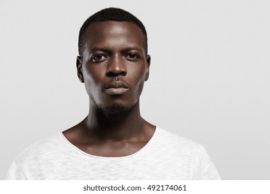 Headshot of attractive serious African student with small beard and moustache, dressed in casual t-shirt, looking at camera with confident and thougthful expression on his face standing at gray wall