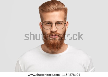 Headshot of attractive serious Caucasian male with thick ginger beard and trendy haircut, dressed in casual white t shirt, looks directly at camera, wears round glasses, isolated over white background