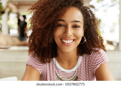 Headshot Of Beautiful Dark Skinned Female With Happy Expression Has Afro Hairstyle, Demonstrates Perfect White Even Teeth, Has Pleased Smile. Stylish Young African American Woman Rests Indoor