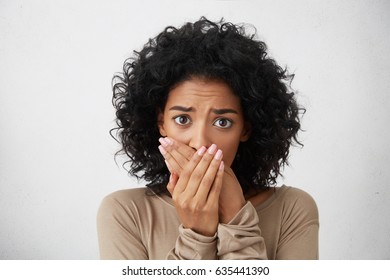 Headshot Of Beautiful Scared Young Dark-skinned European Female With Curly Hairstyle Closing Mouth Not To Scream, Feeling Frightened And Terrified, Her Eyes And Look Full Of Fear And Terror