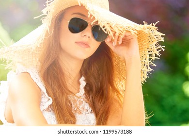 Headshot of beautiful young woman wearing straw hat and sunglasses while sitting outdoor and looking at camera.