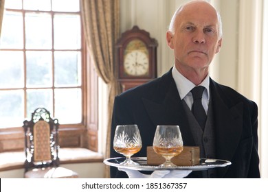 Headshot of a butler wearing a tailcoat standing with a tray containing two glasses of whiskey and a box of cigars