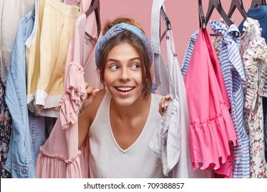 Headshot Of Charming Playful Young European Female Playing Hide And Seek With Little Brother, Hiding Herself Among Lots Of Clothes Hanging On Rack In Her Wardrobe. Shopping And Consumerism Concept