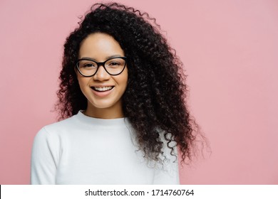 Headshot Of Charming Young Female With Afro Hairstyle, Smiles Pleasantly At Camera, Feels Very Happy, Wears Transparent Glasses And White Jumper, Isolated On Rosy Background. Amused Ethnic Female