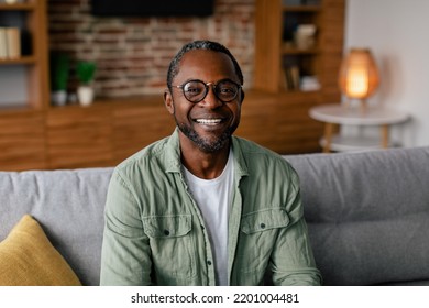 Headshot of cheerful adult african american man in glasses and casual relaxing on sofa and looking at camera in living room interior, close up. Business and work at home during covid-19 quarantine - Powered by Shutterstock