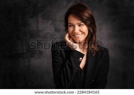 Headshot of a cheerful smiling middle aged woman posing at isolated dark background. Brunette haired female wearing blazer. Copy space. Studio shot.