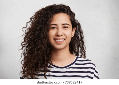 Headshot of curly mixed race beautiful female with smile wears striped sweater, isolated over white background. Caucasian young woman has crisp dark hair and appealing appearance. Beauty concept