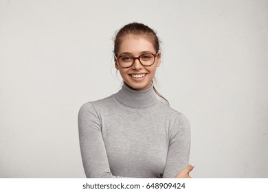 Headshot of cute student girl wearing grey turtleneck sweater and stylish eyeglasses smiling broadly, having carefree and relaxed look while resting at home after morning lectures at college