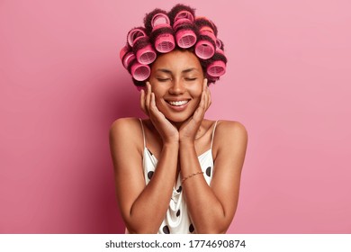 Headshot of dark skinned pleased female model creats perfect hairstyle with hair rollers, touches cheeks with hands, closes eyes and stands bare shouldered against pink background. Beauty concept - Powered by Shutterstock