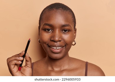 Headshot dark skinned young woman and short hair holds red lip pencil applies makeup wants to look beautiful smiles gently poses bare shoulders isolated over brown background  Beauty concept