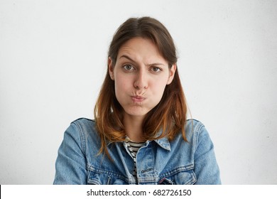 Headshot of displeased European woman blowing her cheeks being upset raising her eyebrows, demonstrating her discontent. Female in jean jacket with sulky expression isolated over white background