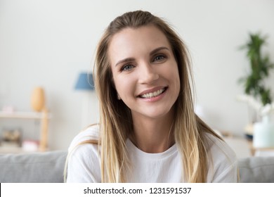 Headshot front portrait of smiling friendly millennial woman sitting alone on couch at home looking talking at camera, attractive casual happy young lady with beautiful face posing indoors