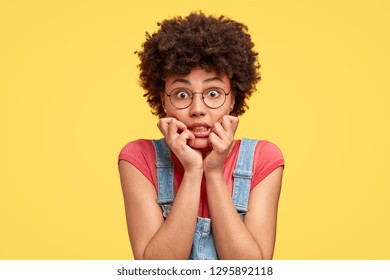 Headshot of frustrated mixed race young woman has crisp hair, bites finger nails with nervous expression, stares with eyes full of fear, wears casual clothes, stands against yellow studio wall