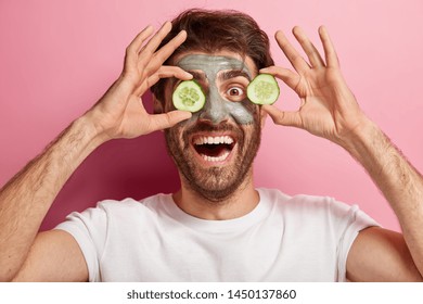 Headshot of funny guy smiles broadly, has clay mask on face, covers eyes with slices of cucumbers, has spa treatments, wears casual white t shirt, poses indoor. Men, beauty, cosmetology concept