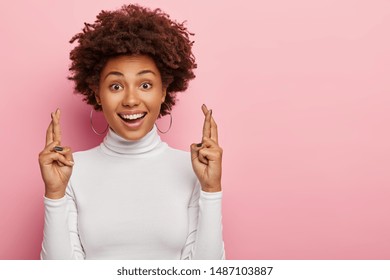 Headshot of glad smiling curly woman crosses fingers with positive expression, believes in good luck and success, looks hopefully straightly at camera, isolated over pink wall with blank space aside