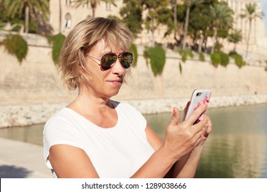 Headshot Of Good-looking Modern Blond Mature Woman Wearing Glasses, Messaging Best Friend Or Husband Via Social Networks, Using Generic Cell Phone, Smiling While Reading Message Or Looking At Photo.