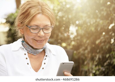 Headshot Of Good-looking Modern Blond Mature Woman Wearing Glasses, Messaging Her Grandson Via Social Networks, Using Her Generic Cell Phone, Smiling While Reading Message Or Looking At Photo