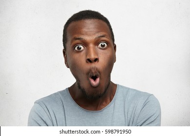 Headshot of goofy surprised bug-eyed young dark-skinned man student wearing casual grey t-shirt staring at camera with shocked look, expressing astonishment and shock, screaming "Omg" or "Wow"