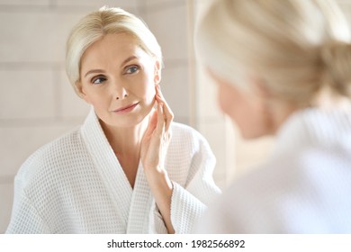 Headshot of gorgeous mid age adult 50 years old blonde woman standing in bathroom after shower touching face, looking at reflection in mirror doing morning beauty routine. Older skin care concept. - Shutterstock ID 1982566892