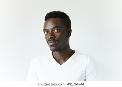 Headshot of handsome young African man wearing white T-shirt, looking at the camera, posing isolated against white concrete wall background with copy space for your information or advertising content