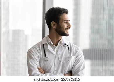 Headshot happy ambitious doctor young expert of modern clinic stand in confident pose look at window with shiny smile. Successful man medic think about professional growth enjoy saving patients health