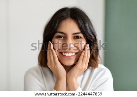 Headshot of happy smiling attractive young arabic lady in white homewear touching her cheeks, enjoying silky skin after using luxury organic premium beauty face care products, closeup
