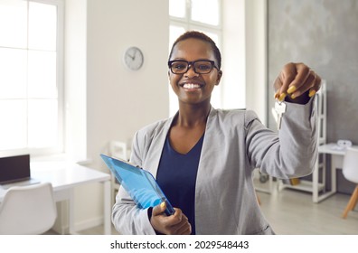 Headshot Of Happy Smiling Realtor Or Real Estate Agent Showing Key To New House. Beautiful Short Haired Black Lady In Suit And Glasses Standing In Office, Holding Clipboard And Keys, Looking At Camera