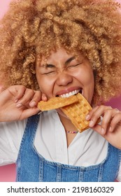 Headshot Of Hungry Curly Woman Bites Appetizing Handmade Waffles Keeps Eyes Closed Wears White Shirt And Denim Sarafan Enjoys Favorite Dessert Poses Indoor. Unhealthy Snack Harmful Nutrition