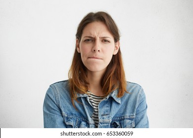 Headshot of indecisive confused young European woman in denim wear pursuing lips, her look expressing doubt and uncertainty as she has to come up with best solution while dealing with problem