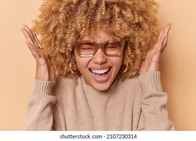 Headshot Of Overjoyed Happy Curly Woman Raises Hands Over Head And Exclaims Joyfully Wears Spectacles And Cashmere Jumper Poses Against Brown Background Reacts On Amazing News. Emotions Concept