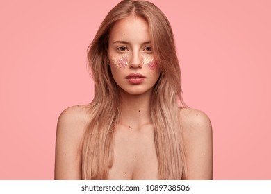 Headshot of pleasant looking female with beautiful art makeup, glitters on face, poses lalf nude, has long straight hair, poses against pink studio background. Lovely woman has sparkles on cheeks