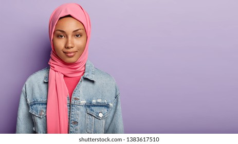 Headshot Of Pleased Lovely Muslim Lady Has Gentle Smile, Healthy Skin, Wrapped In Silk Pink Hijab, Has Natural Beauty, Models Over Purple Background With Blank Space For Text. People And Religion