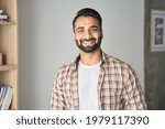 Headshot portrait of attractive confident indian Hispanic man with toothy smile looking at camera at modern living room. Latin businessman posing in casual stylish look at home office.