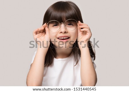 Headshot portrait of cute little preschooler girl isolated on grey studio background wear glasses look at camera, small child try spectacles at opticians, kid eyesight correction treatment concept